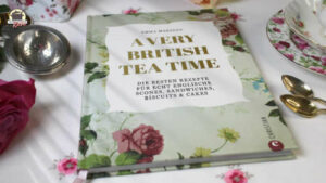 A very british Teat Time liegt auf einer mit Rosen bestickten Tischdecke. Daneben liegt eine rosafarbene Rose sowie zwei silberne Löffel und ein silbernes Teesieb.