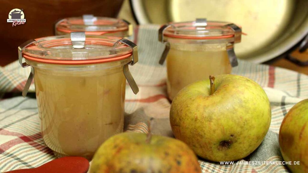 Frische Äpfel und drei Gläser mit Apfelmarmelade stehen auf einem kariertem Tuch.