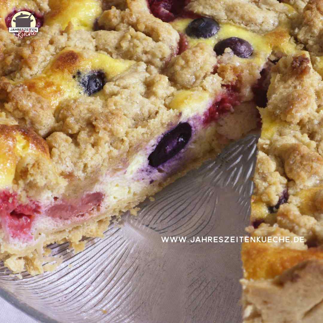 Auf einem Glasteller ist ein angeschnittener Beeren-Käsekuchen mit Streusel