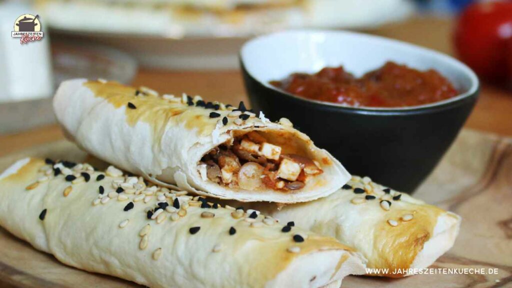 Ein aufgeschnittenes Börek-Röllchen liegt auf zwei weiteren Börek. Im Hintergrund steht eine Schälchen mit Chutney.