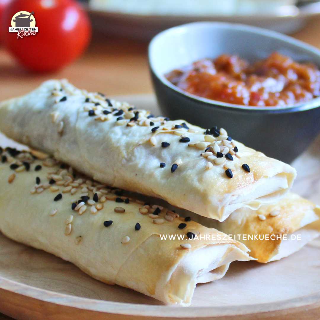 Drei Sigara Börek mit Linsenfüllung liegen auf einem Holzteller. Im Hintergrund ist ein Schälchen mit Tomaten-Chili-Chutney und eine Tomate.