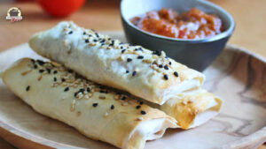 Drei Sigara Börek mit Linsenfüllung liegen auf einem Holzteller. Daneben steht eine Schälchen mit Tomaten-Chili-Chuntey.