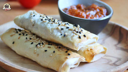 Drei Sigara Börek mit Linsenfüllung liegen auf einem Holzteller. Daneben steht eine Schälchen mit Tomaten-Chili-Chuntey.