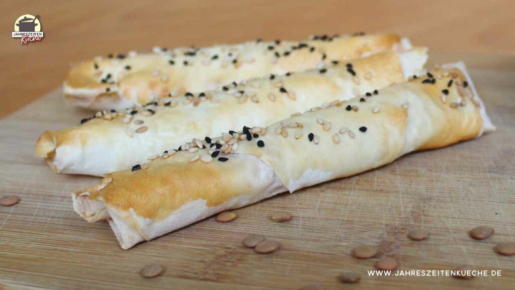 Drei Sigara Börek mit Linsenfüllung liegen auf einem Holzbrett. Darauf sind getrocknete Linsen verstreut.