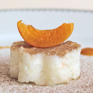 Eine Aprikosenspalte liegt auf einer Portion Buttermilchreis, die Zimt-Zucker bestreut ist.