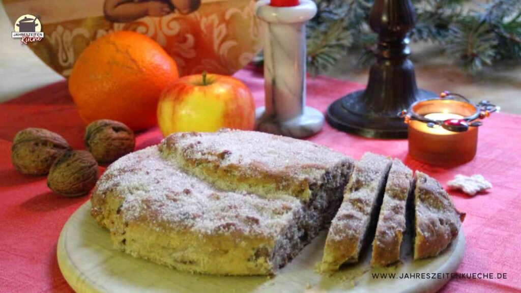 Christstollen mit Marzipan und Datteln auf einem Holzbrett. Im Hintergrund sind Orange, Apfel, Kerze
