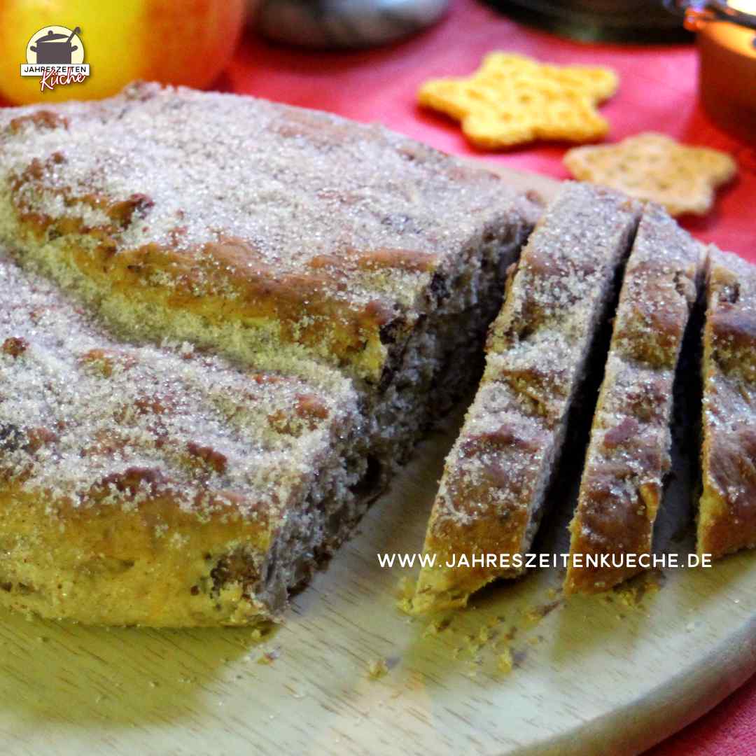 Auf einem Holzbrett liegt ein angeschnittener Christstollen mit Marzipan und Datteln. Im Hintergrund sind Orange, Apfel, Kerze