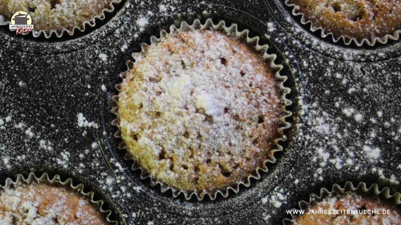 Ein mit Puderzucker bestreuter Muffin ist in der Mitte von einem schwarzen Muffinblech.