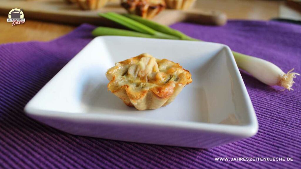 Eine Frühlingszwiebel-Pastete liegt auf einem weißen Teller. Im Hintergrund liegen weitere Pasteten auf einem Holzbrett