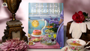 Das Kochbuch Genießen wie bei den Bridgertons ist eingerahmt von einem alten Kerzenleuchter und einem Stapel Bücher, auf denen weiße Handschuhe und eine rote Rose liegen.