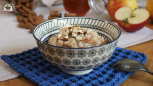 Ein Schälchen mit Habermus auf einem blauen Deckchen. Im Hintergrund liegen Äpfel, Honig und Mandeln.