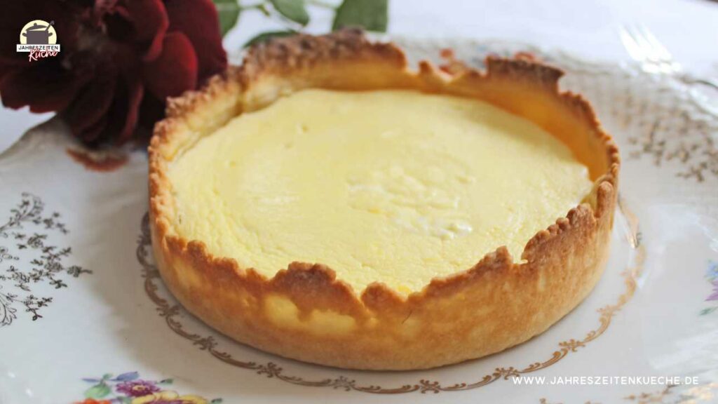 Ein Käsekuchen in Singlegröße auf einem Teller mit Blumenmuster. Im Hintergrund liegt eine dunkelrote Rose.