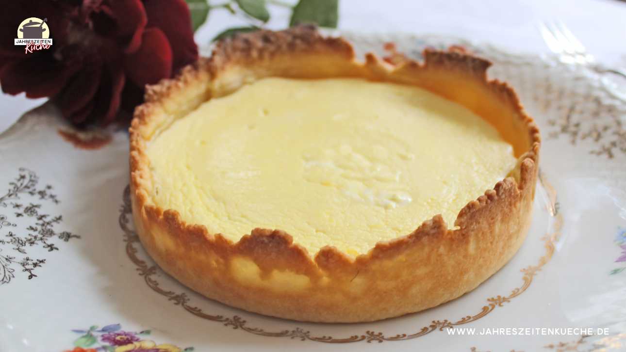 Ein Käsekuchen in Singlegröße auf einem Teller mit Blumenmuster. Im Hintergrund liegt eine dunkelrote Rose.