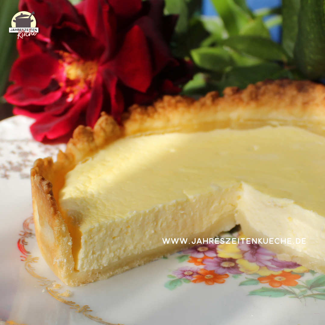 Auf einem Teller liegt ein kleiner Käsekuchen, von dem ein Viertel fehlt. Dahinter liegt eine dunkelrote Rose