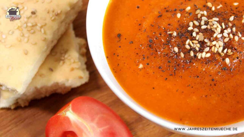 Eine weiße Suppenschale mit Karotten-Tomaten-Suppe. Links daneben liegt eine Tomaten und Lavashbrot.