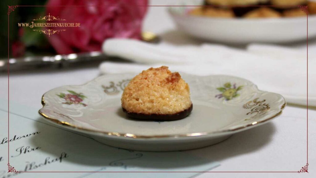 Eine einzelne Kokosmakrone steht auf einem Portionsteller. Im Hintergrund steht ein Teller mit vielen Kokosmakronen