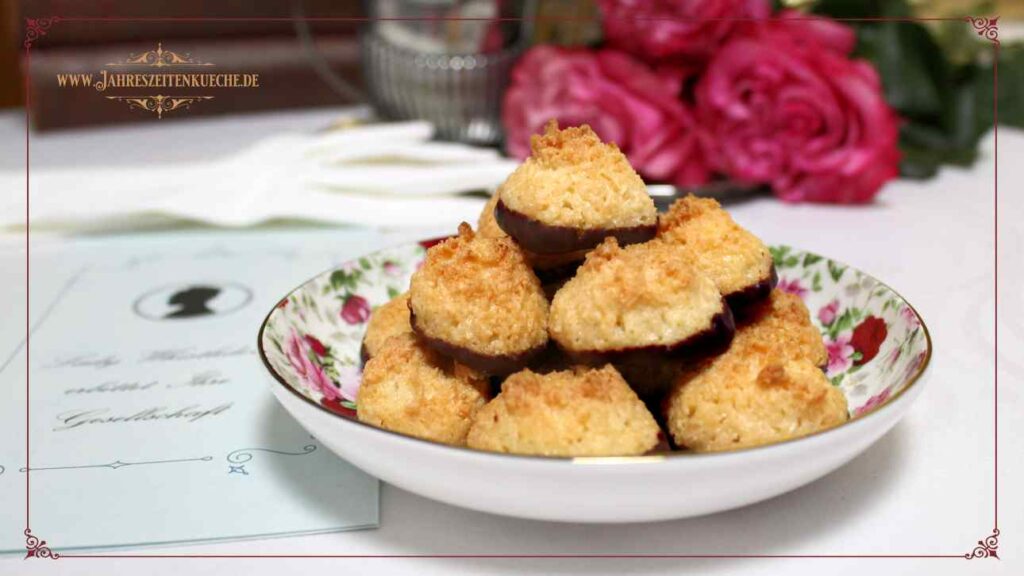 Ein Pyramide aus Kokosmakronen ist auf einem Telelrchen. Links daneben liegt eine Einladungskarte von Lady Whistledown und im Hintergrund rote Rosen