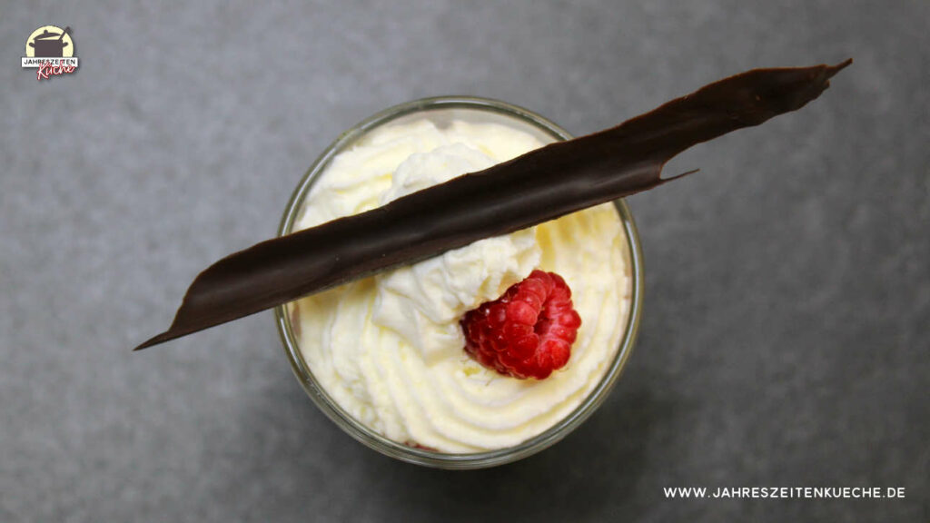 Auf einer Schiefer-Platte steht ein Glas mit Mandelcreme, Sahne und Himbeere, Quer darüber liegt ein langer Schokoladenspan.