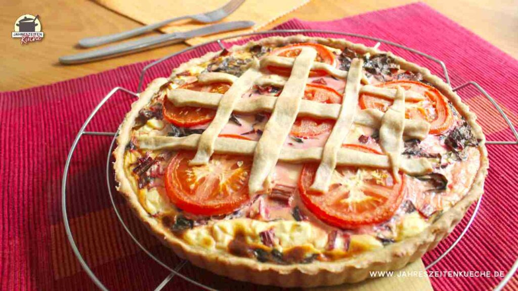 Mangoldquiche mit Tomatenscheiben auf einem Küchengitter. Im Hintergrund liegen Messer und Gabel.