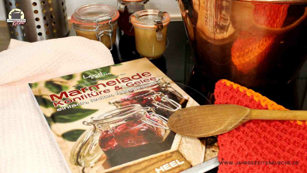 Neben dem Buch Marmelade, Konfitüre & Gelee steht ein Kupfertopf, davor liegt ein Kochlöffel und im Hintergrund stehen kleine Marmeladengläser