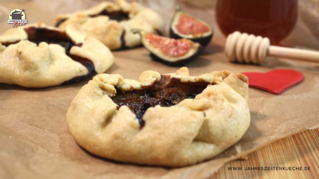 Drei kleine Mini-Galettes liegen auf einem Stück Backpapier. Im Hintergrund liegen Feigen und ein Honiglöffel.