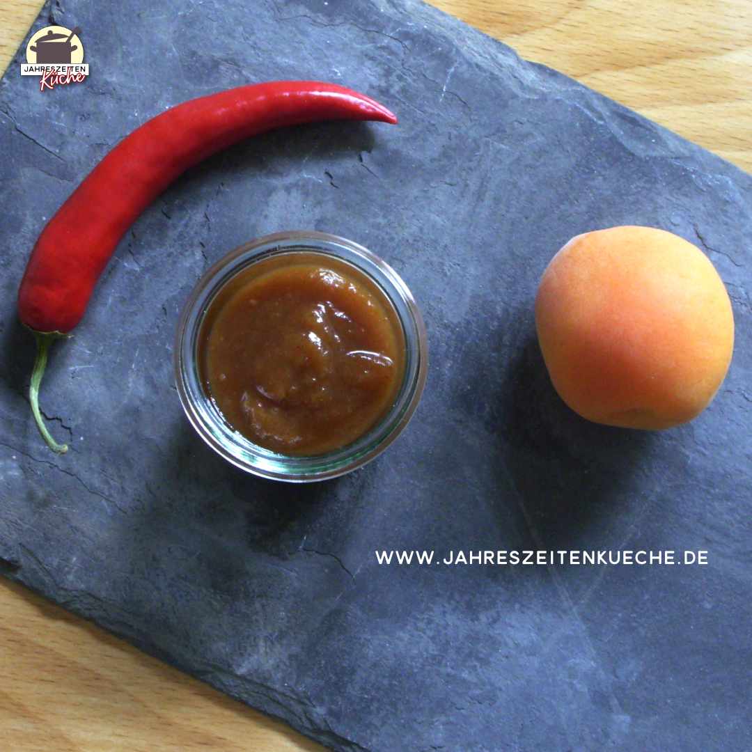 Ein kleines Glas Mirabellen-Aprikosen-Chutney steht auf einer Schieferplatte. Daneben liegen eine rote Chilischote und eine Aprikose