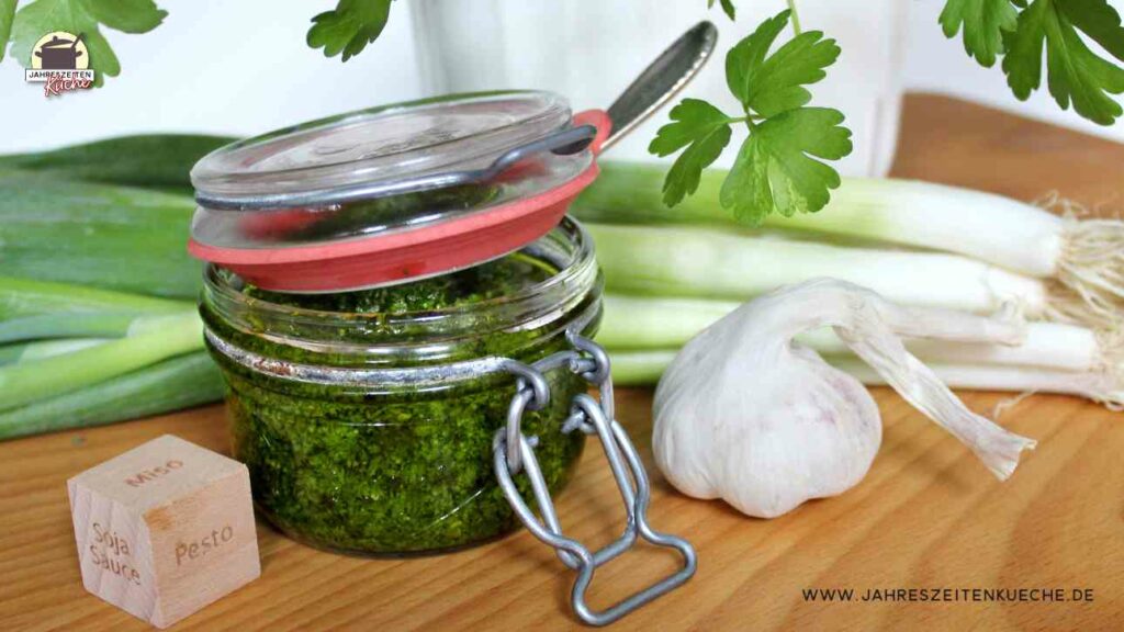 In einem Drahtbügelglas mit Petersilienpesto steckt ein Löffel. Daneben liegt Knoblauch und dahinter Frühlingszwiebeln.