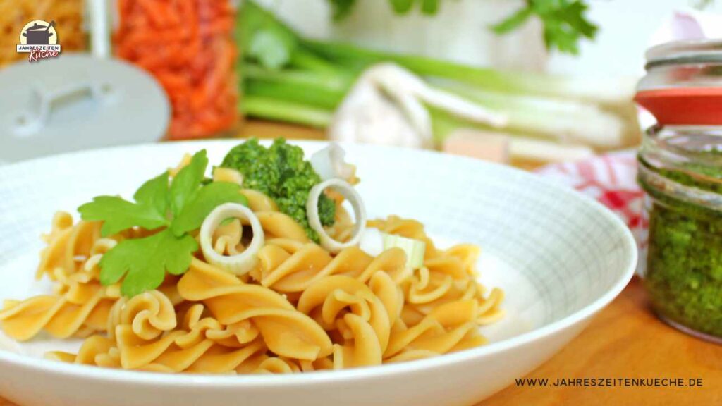 Spirelli-Nudeln mit Persilienpesto auf einem Teller. Im Hintergrund sind Gläser mit roten und gelben Nudeln, Knoblauch, Frühlingszwiebeln und ein Drahtbügelglas mit Petersilienpesto.