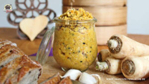 Ein Glas Petersilienwurzel-Aufstrich steht auf einem Holzbrett. Daneben liegt ein aufgeschnittenes Brot.