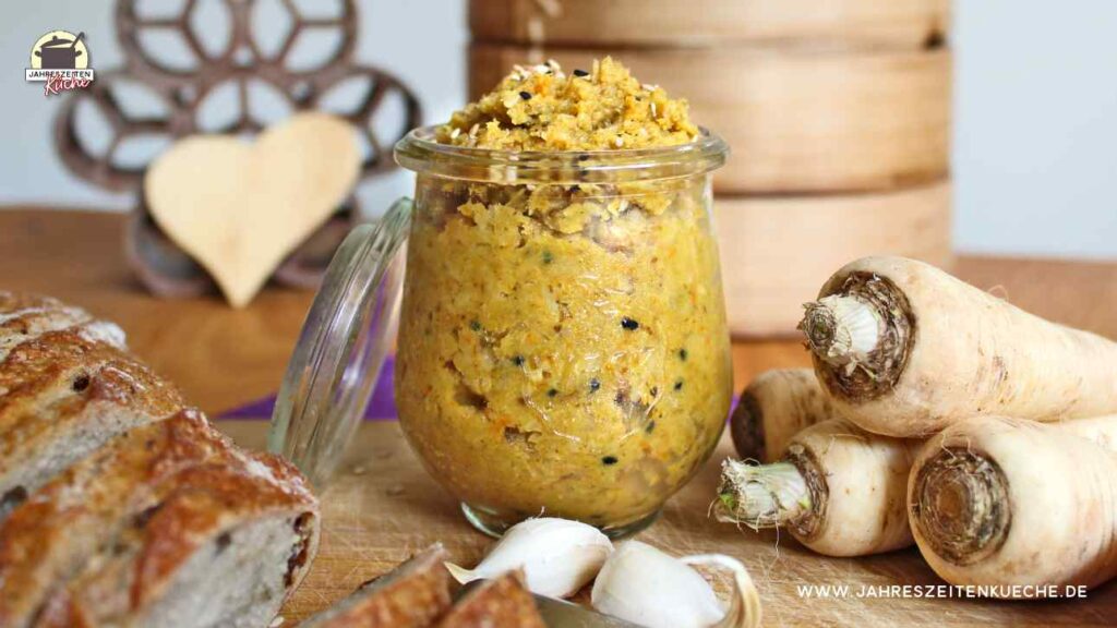 Ein Glas Petersilienwurzel-Aufstrich steht auf einem Holzbrett. Daneben liegt ein aufgeschnittenes Brot.