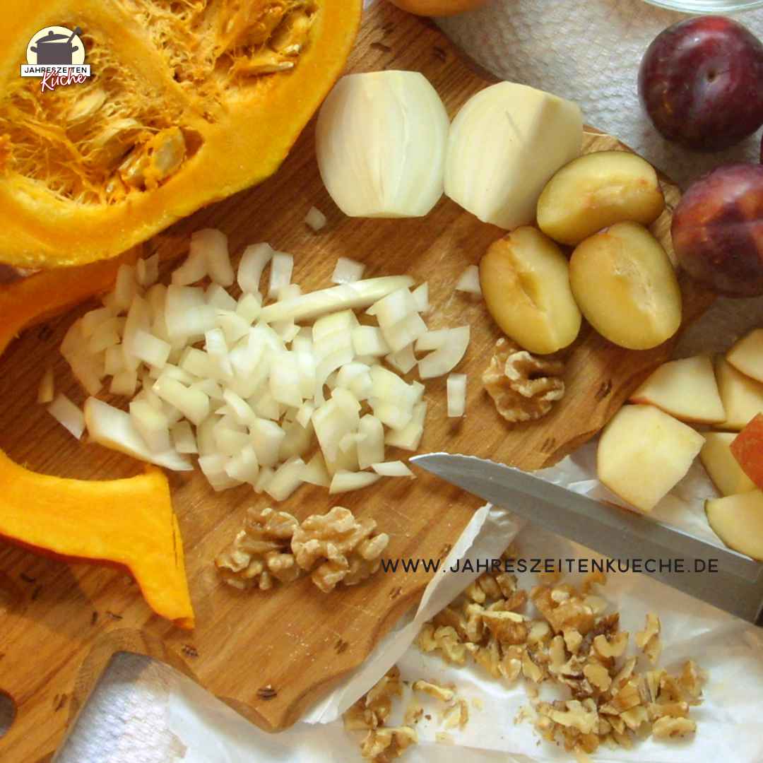 Hokkaido-Kürbis, Zwiebeln, Walnüssen, Pflaumen, Äpfel und ein Messer liegen auf einem Holzbrett