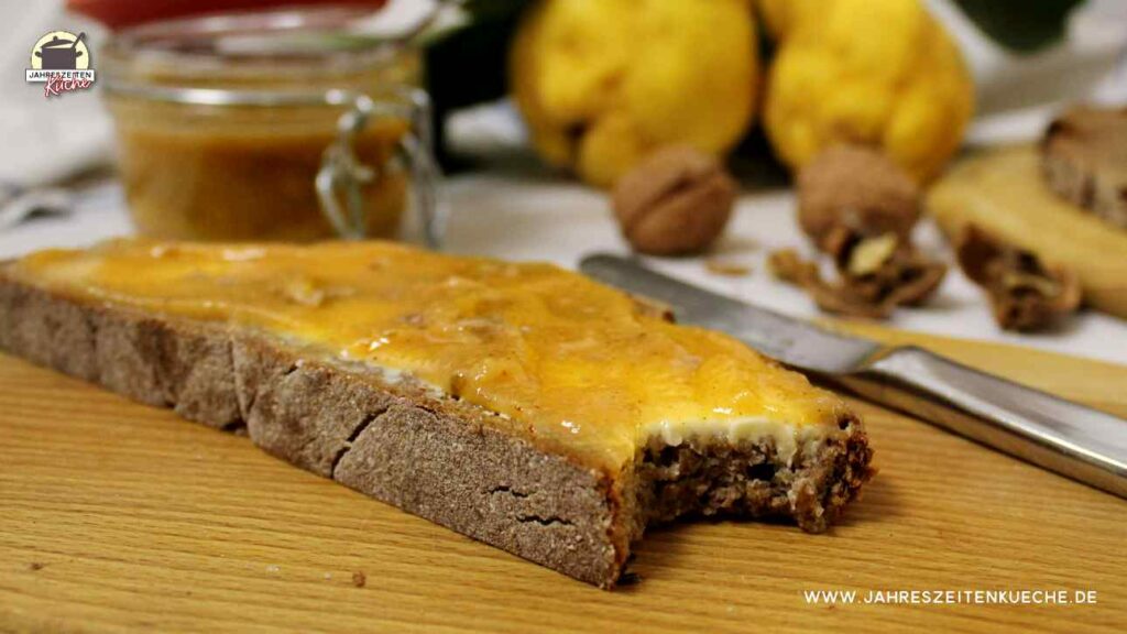 Eine angebissene Scheibe Brot mit Quittenmarmelade. Im Hintergrund liegen Quitten und Walnüsse.
