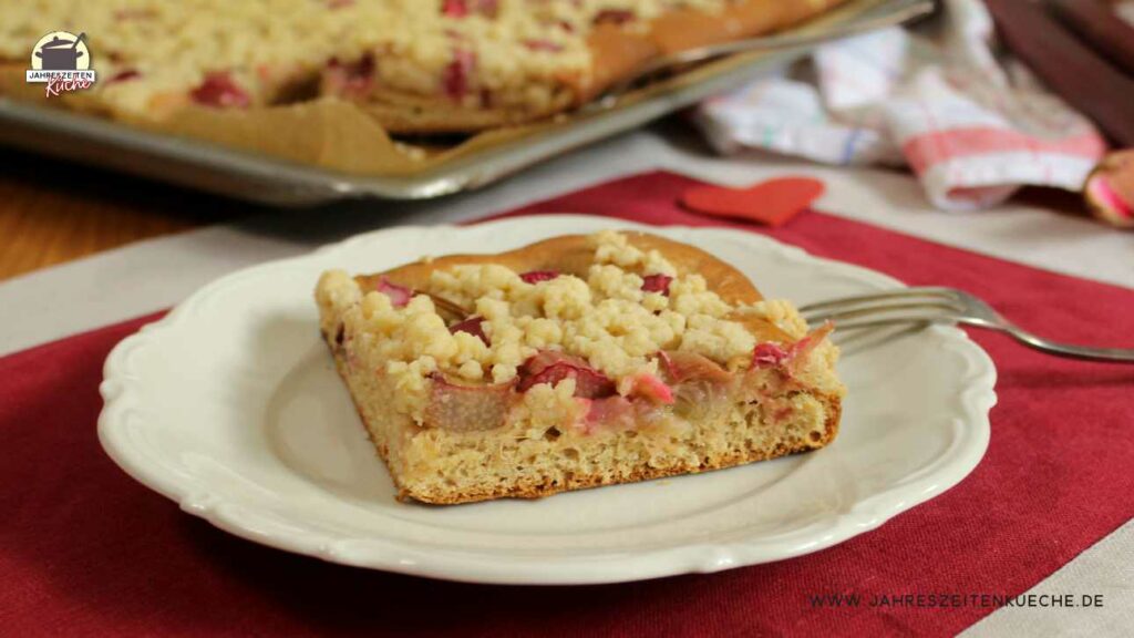 Auf einem weißen Teller liegt ein Stück Rhabarberkuchen. Im Hintergrund ist der Blechkuchen zu sehen