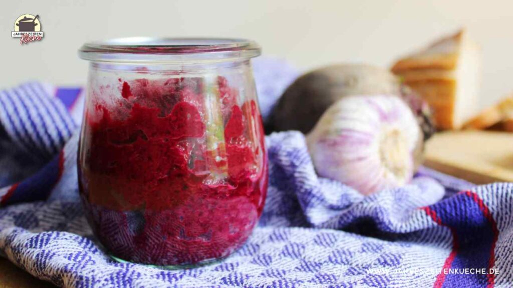 Auf einem blauen Tuch steht ein Glas Rote Bete-Aufstrich. Im Hintergrund Knoblauch und Rote Bete.