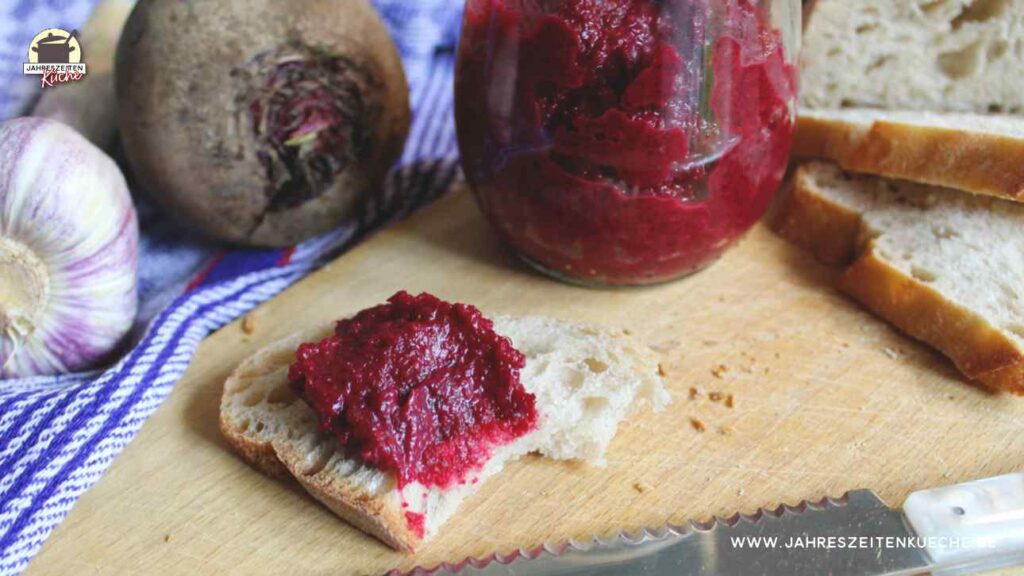 Ein Stück Brot mit Rote Bete-Aufstrich. Im Hintergrund ein Glas mit Rote Beete-Aufstrich.