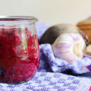 Auf einem blaukarierten Tuch steht ein Glas Rote-Bete-Aufstrich, Knoblauch und Rote Bete-Knollen