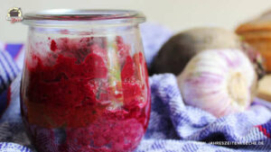 Auf einem Holzbrett steht ein Glas Rote-Bete-Aufstrich neben ein paar Scheiben Brot. Danben liegen Knoblauch und Rote Bete-Knollen.