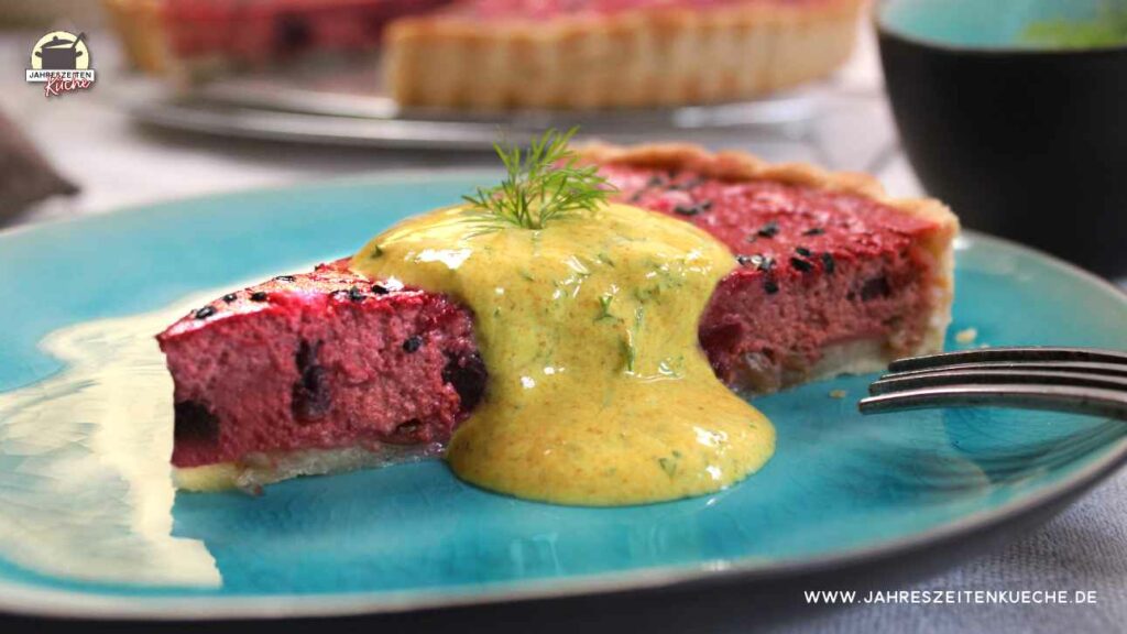 Ein Stück Rote-Bete-Quiche mit Feta liegt auf einem türkisfarbenen Teller. Im Hintergrund ist die angeschnittene Quiche zu sehen.
