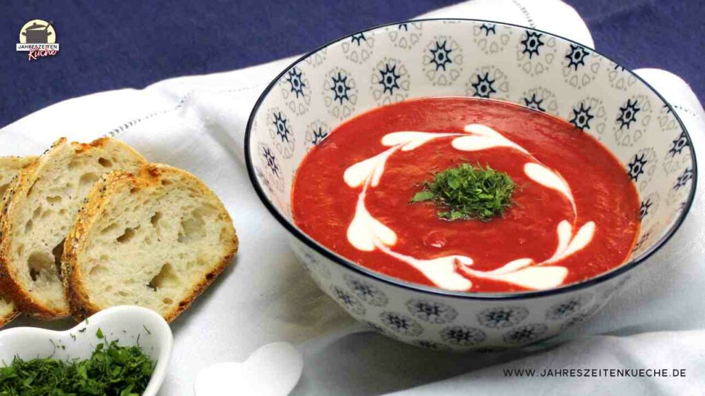 Kartoffelsuppe mit Rote Beete und Chili ist in einer Schüssel, die auf einem weißen Tischtuch steht. Links daneben liegen drei Scheiben Brot.