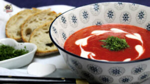 Rote-Beete-Kartoffelsuppe mit frischen Kräutern und Saure Sahne in einer Schüssel. Im Hintergrund sind drei Scheiben Brot.