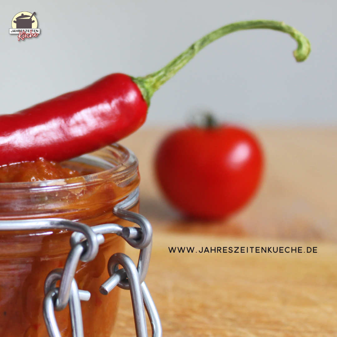 Auf einem Glas mit Tomaten-Chili-Chutney liegt eine Chilischote, im Hintergrund eine Tomate.