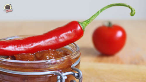 Eine Chilischote liegt auf einem Glas mit Tomaten-Chutney. Im Hintergrund ist eine Tomate zu sehen.