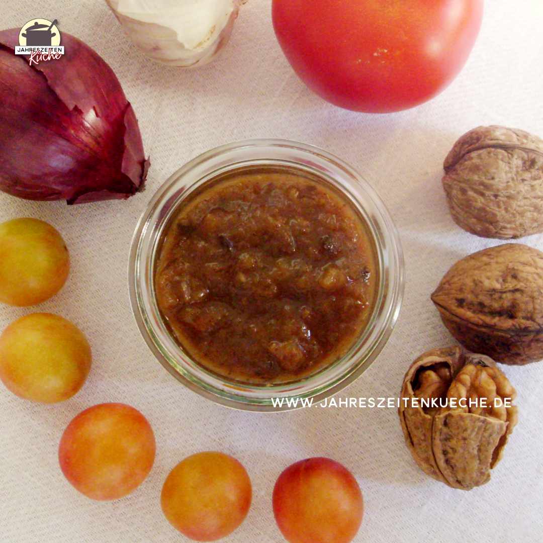 In einem Kreis aus roter Zwiebel, Knoblauch, Tomate, Walnüssen und Wildplaumen steht eine Glas Tomaten-Wildpflaumen-Chutney