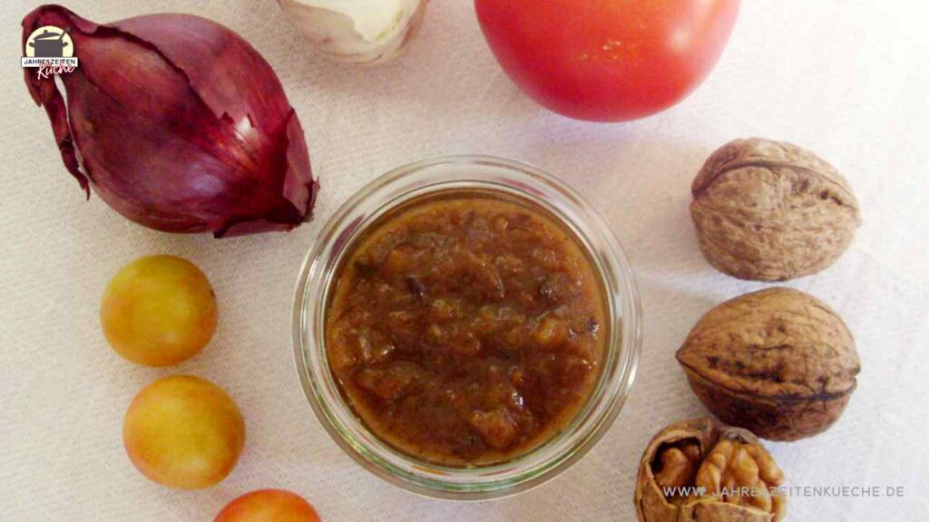 Um ein Glas Tomaten-Wildpflaumen-Chutney liegen Knoblauch, rote Zwiebel, Wildpflaumen, Tomaten und Walnüsse