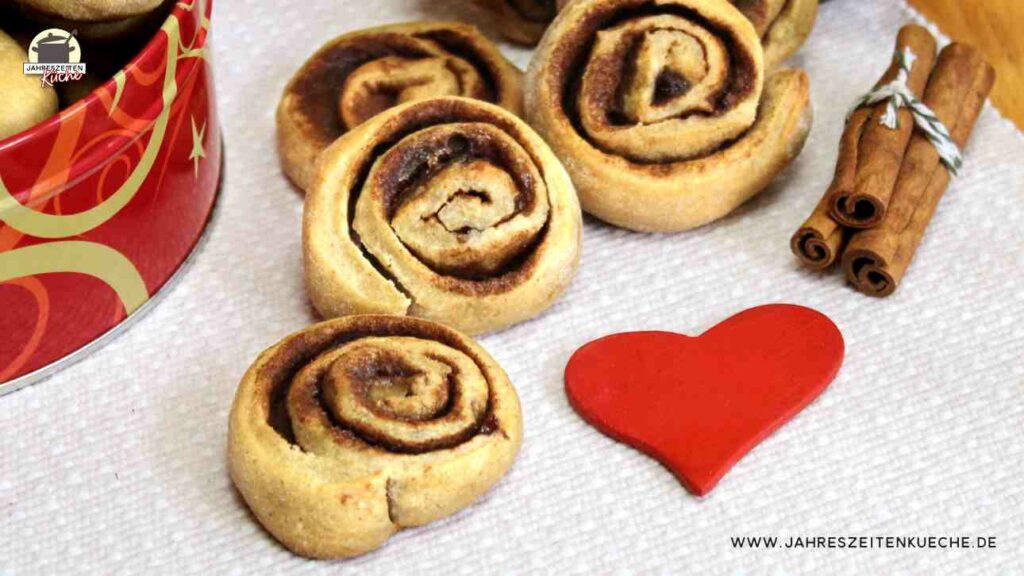 Neben einer roten Dose liegen mehrere Zimtschnecken, Zimtstangen und ganz rechts ein rote Herz.
