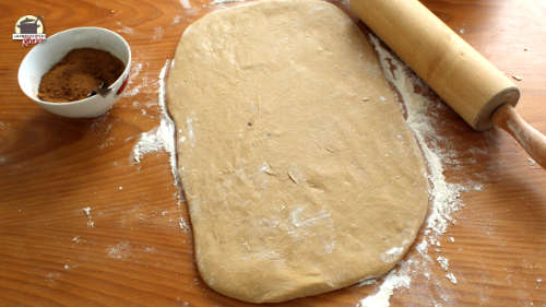 Rechteckig ausgerollter Zimtschnecken-Teig liegt auf einem Holztisch. Rechts daneben eine Teigrolle