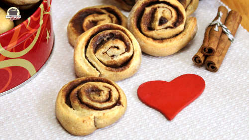 Frisch gebackene Zimtschnecken liegen auf hellem Stoff. Rechts daneben sind Zimtstangen und im Vordergrund ein rotes Herz.
