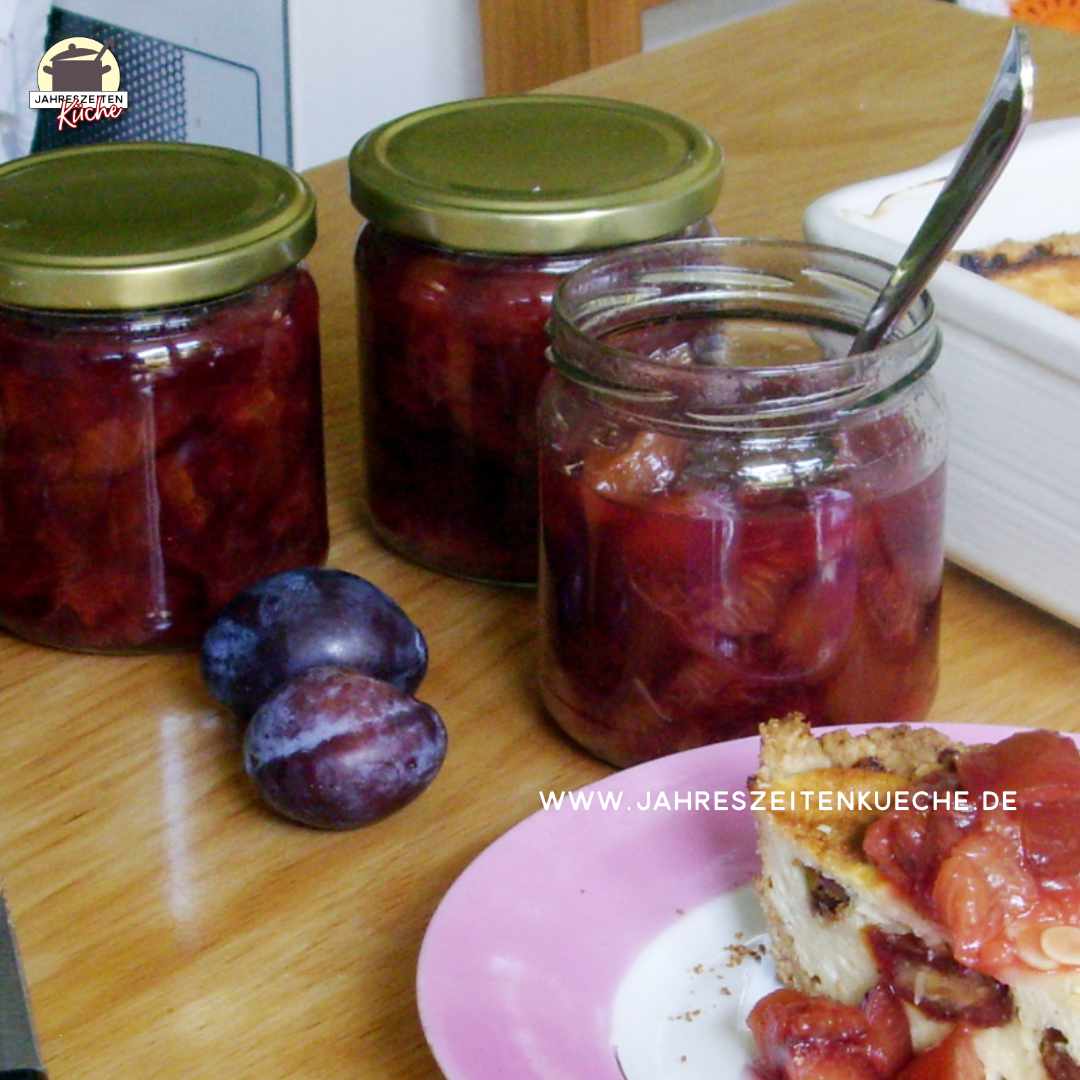 Auf einem Holztisch stehen Gläser Zwetschgenkompott. Davor steht ein Stück Käsekuchen.