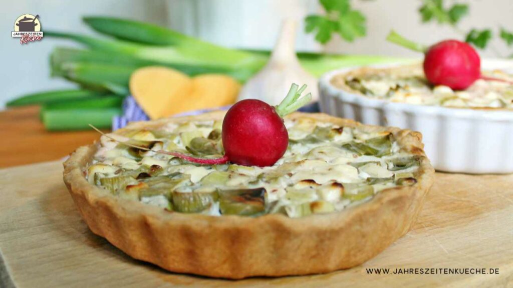 Zwiebelquiche mit einem Radieschen garniert auf einem Holzbrett. Im Hintergrund ist eine zweite Quiche in einer weißen Keramikform.