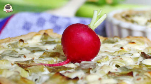 Zwiebelquiche mit einem Radieschen garniert auf einem Holzbrett. Im Hintergrund ist eine zweite Quiche in einer weißen Keramikform.